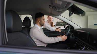 Genç bir adam bir galeride yeni bir otomobilin içinde oturmuş, kadın bir satış danışmanı satın alma kararı için bilgi verirken ve yardım ederken, içişleri ve direksiyon simidini inceliyor.