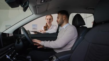 Profesyonel kadın araba satıcısı yeni bir aracın içinde oturan bir erkekle konuşuyor, galeride test sürüşü sırasında arabanın içini ve özelliklerini gösteriyor.