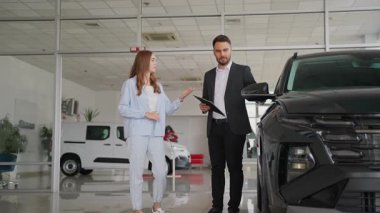 Profesyonel bir araba satıcısı, geniş ve parlak bir galeride, ilgilenen genç bir bayana, modern siyah bir kesişme noktası sunarak, ona bir araç seçmesinde yardımcı oluyor.