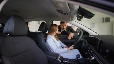 Genç bir kadına yeni bir araç sunan bilgili bir araba satıcısı, modern bir galeride sürücü koltuğunda otururken gösterge tablosunun özelliklerini ve teknolojisini ön plana çıkarıyor.