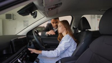 Genç bir kadın galeride yeni bir arabanın şoför koltuğunda otururken profesyonel bir erkek araba acentesi durumu açıklıyor.