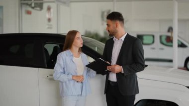 Profesyonel erkek danışman genç bir kadına araç özelliklerini ve kontrat detaylarını açıklıyor. Müşteri, modern bir araba galerisinde yeni bir otomobil seçerken dikkatle dinliyor.