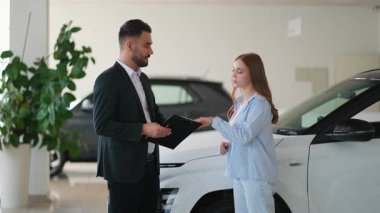 Yeni bir araba için satış anlaşması imzalayan genç bir kadın başarılı bir alışveriş sonrasında modern bir otomobil bayiliğinde satıcılarla el sıkışıyor ve anlaşmayı tamamlıyor.