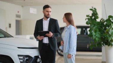 Modern bir bayilikte genç bir bayana yeni bir beyaz otomobil gösteren profesyonel erkek danışman, araç özelliklerini tartışıyor ve ihtiyaçları için doğru arabayı seçmesine yardım ediyor.