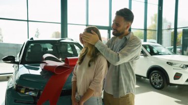 Sevgi dolu koca, mutlu karısına gözlerini kapayarak ve büyük kırmızı bir fiyonkla dekore edilmiş yeni bir arabayı göstererek modern araba galerisinde duygusal ve neşeli bir öpücüğe yol açarak sürpriz yapar.