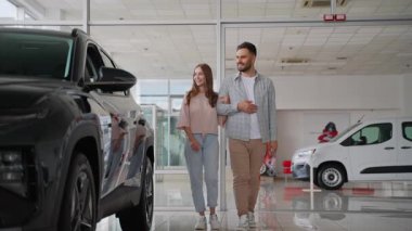 Modern bir araba galerisinde birlikte yürüyen mutlu genç çift, yeni araçlara bakıyor ve alışverişlerini tartışıyorlar. Kadın, kararlarını verirken siyah bir cipin yanına dokunuyor.