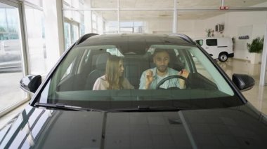 Yeni bir galeride yeni arabalarında oturan mutlu genç çift, satın almaları ile ilgili saf neşe ve heyecanı dile getiriyorlar, modern bir aile arabası almanın başarısını kutluyorlar.