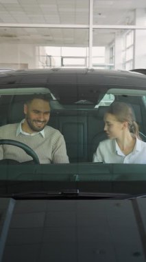 Genç bir çift, bir bayide yeni bir modern arabada oturuyor, satın alma kararı vermeden önce kadın satış müdürüyle konuşuyor ve otomobilin içini inceliyor.