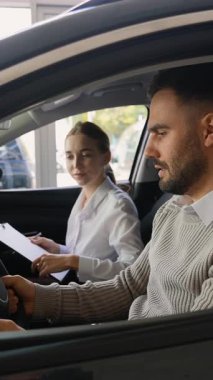 Yeni bir aracın sürücü koltuğunda oturan yakışıklı erkek müşteri, iç mekanı inceliyor ve modern bir araba galerisinde profesyonel bir kadın satış müdürüyle konuşuyor.