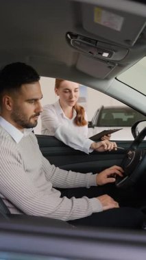 Araba galerisinde çalışan profesyonel bir kadın satış temsilcisi yeni bir otomobil alımı için erkek bir müşteriye danışıyor. Direksiyon başında otururken ona iç mekan özelliklerini ve kontrollerini gösteriyor.