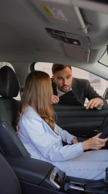 Yeni bir arabanın sürücü koltuğunda oturan genç kadın, modern bir galeride arabaların özelliklerini ve sözleşme detaylarını açıklayan erkek araba satıcısını dinliyor.