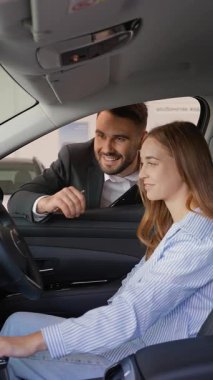 Profesyonel bir araba satıcısı, genç bir bayana galeride yeni bir araç için danışıyor, ona içeride direksiyon başında otururken arabanın özelliklerini gösteriyor.