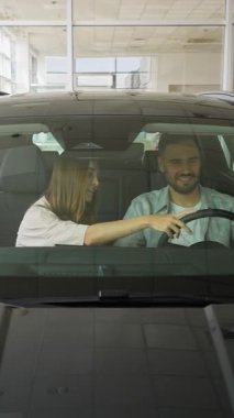 Bir araba galerisinde modern bir araçta oturan neşeli genç çift, satın almadan önce içeriyi ve özellikleri büyük bir coşkuyla tartışıyor, mutlu bir satın alma deneyimi sergiliyorlar.