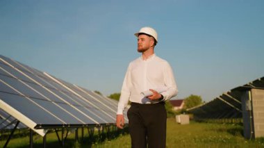 Güneş enerjisi santralinde yürürken elinde planları tutan bir kasketin içindeki erkek enerji mühendisi. Sürdürülebilir ve yenilenebilir enerji kavramı