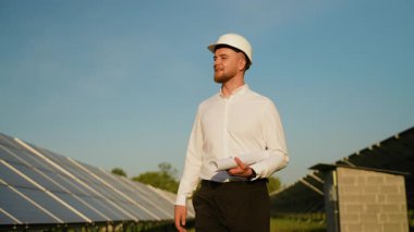 Güneş paneli çiftliğinde yürürken elinde planlar tutan sağlam şapkalı kendinden emin bir erkek mühendis. Yenilenebilir enerji ve yeşil güç kavramı