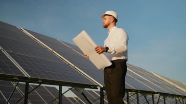 Erkek güneş enerjisi mühendisi elinde planlarla kırsal kesimdeki bir elektrik santralinin güneş paneli kurulumunu kontrol ediyor.