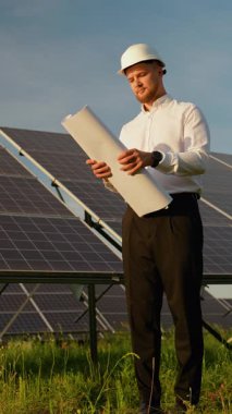 Beyaz şapkalı kendinden emin erkek mühendis teknik bir çizimi inceliyor. Gün batımında güneş panellerinin önünde bir tarlada duruyor.