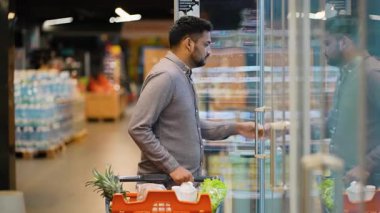 Young man with a shopping cart choosing a carton of eggs from the refrigerated section of a grocery store