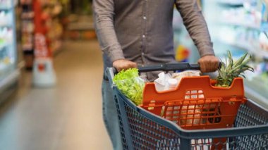 Tanınmayan erkek müşteri, modern bir süpermarketin koridorunda içi taze ürün ve sebze dolu bir alışveriş arabasını itiyor ve günlük sağlıklı ve organik gıda alımlarını yapıyor.
