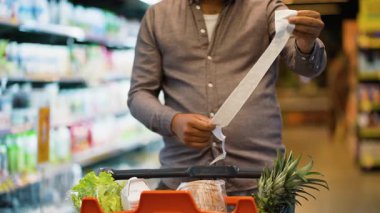 Afro-Amerikan bir adamın, marketteki harcamalarını kontrol ederken, içi taze yiyecek, ananas, ekmek ve marul dolu bir market arabasıyla uzun bir alışveriş fişini dikkatlice incelemesine dikkat edin.