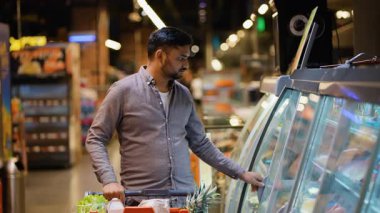 Alışveriş arabalı yakışıklı Hintli adam büyük bir markette soğutuculu vitrinden taze ürünler seçiyor sağlıklı bir yemek için düşünceli seçimler yapıyor.