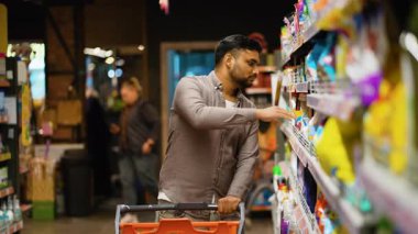 Alışveriş arabalı genç Hintli bir adam süpermarket koridorunda kimyasalları ve çamaşır deterjanını seçiyor, satın almadan önce ürün etiketini dikkatlice okuyor.