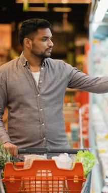 Genç Hintli bir adam marketin koridorunda bir alışveriş arabasını itiyor, buzdolabı bölümünden dikkatlice bir süt ürünü seçiyor ve sepetine koyuyor.