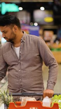 Odaklanmış genç Hintli bir adam ürünleri dikkatle raflardan seçiyor ve onları alışveriş arabasına koyup modern bir marketin veya süpermarketin koridorlarına bakıyor.