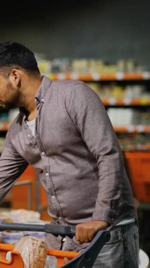 Genç adam bir süpermarketin koridorunda bir alışveriş arabasını itiyor, haftalık market alışverişi sırasında sepetine eklemek için meyve bölümünden olgun bir ananas seçiyor.