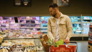 Yakışıklı sakallı adam marketteki soğutuculu vitrinden peynir seçiyor ve alıyor. Malı arabasına koyuyor.