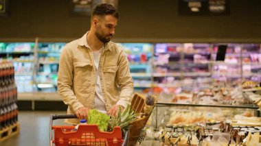 Odaklanmış bir adam alışveriş arabasını itiyor ve modern bir marketteki vitrinden peyniri dikkatlice seçiyor. Sağlıklı yiyecek seçimi kavramı