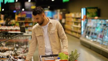 Alışveriş arabalı yakışıklı bir erkek müşteri marketin şarküteri bölümündeki dondurulmuş bir vitrinden dilimlenmiş et seçiyor.