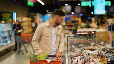 Yakışıklı adam market alışverişi yapıyor, modern bir süpermarket koridorunda soğutulmuş et ve sosis seçiyor.