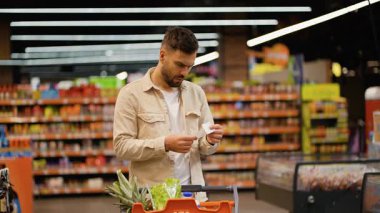 Alışveriş listesini gözden geçiren konsantre genç bir adam, bir market koridorunda, raflarla çevrili bir tramvayla dikilirken