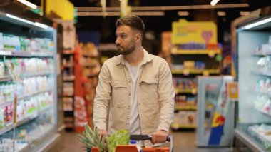 Market koridorunda alışveriş arabasıyla yürüyen düşünceli bir genç adam. Modern bir süpermarkette taze ürünler seçen bir erkek müşteri.