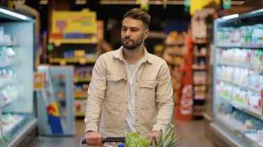 Kendine güveni tam genç bir adam marketin koridorunda yürürken bir alışveriş arabasını taze ürünlerle doldurup doğrudan kameraya bakıyor.