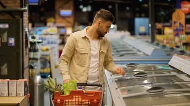 Alışveriş arabalı genç yakışıklı adam modern bir süpermarkette dondurucudan donmuş ürünleri seçiyor. Sağlıklı yiyecekler için market alışverişi
