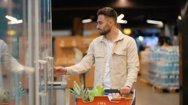 Alışveriş arabalı genç yakışıklı adam modern bir markette ya da süpermarkette dondurulmuş vitrinden taze yumurta seçiyor.