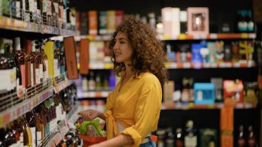 Alışveriş sepeti olan kıvırcık saçlı genç bir kadın marketin alkol reyonundan bir şişe gül şarabı seçiyor.