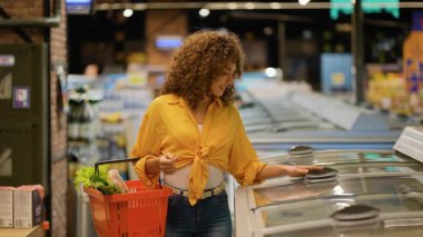 Kıvırcık saçlı genç bir kadın buzdolabından taze somon paketi seçiyor. Müşteri marketten deniz ürünleri seçiyor