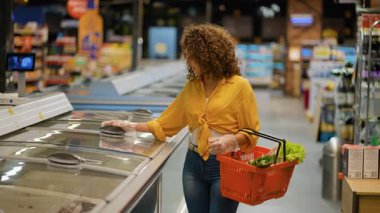 Kıvırcık saçlı genç bir kadın markette dondurucudan donmuş et topluyor. Kadın müşteri günlük alışverişini yapıyor.