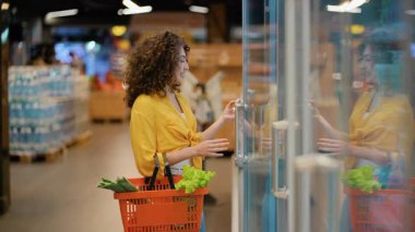 Alışveriş sepeti olan güzel kıvırcık kadın marketteki buzdolabından taze bıldırcın yumurtası seçiyor ve mutlu bir şekilde gülümsüyor.