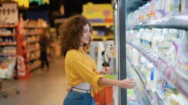 Genç kıvırcık saçlı kadın marketin mandıra bölümündeki soğutuculu raftan bir şişe sütü dikkatlice seçiyor.