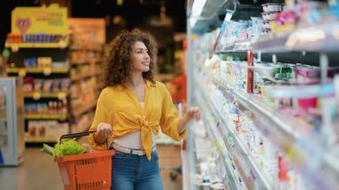 Alışveriş sepeti ile güzel kıvırcık saçlı kadın buzdolabından yoğurt seçiyor. Gitmeden önce etiketi dikkatlice okuyor.