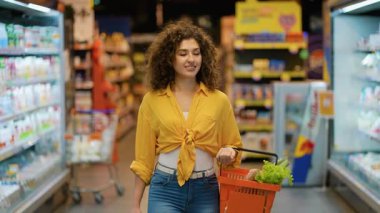 Genç kıvırcık saçlı bir kadın markette yürürken elinde bir sepet dolusu taze sebze ve diğer sağlıklı gıda ürünlerini taşıyor.