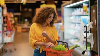 Genç kıvırcık saçlı kadın alışveriş listesini kontrol ederken elinde bir sepet dolusu taze sebzeyle modern bir marketin koridorunda duruyor.