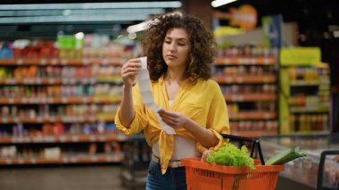 Bir markette elinde alışveriş sepeti olan mutsuz genç bir kadın uzun bir fişe bakıyor. Fiyatlar için endişelenen kadın müşteriler şok oldu ve strese girdi