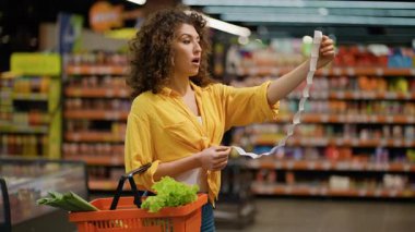 Markette elinde taze sebze dolu bir sepetle duran genç bir kadın, çok uzun bir faturaya şoke olmuş bir şekilde bakıyor.