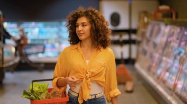 Kıvırcık saçlı çekici genç bir kadın elinde taze sebzelerle kırmızı bir sepet tutuyor. Bir süpermarket koridorunda yürüyor ve ürünleri seçiyor.