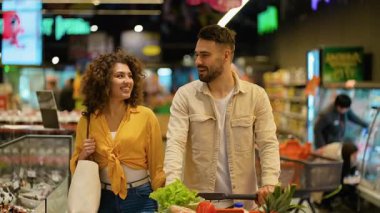 Süpermarket koridorunda yürüyen neşeli bir çift, ürünleri seçiyor ve konuşuyor. Taze sebze dolu bir alışveriş arabasını iten adam.